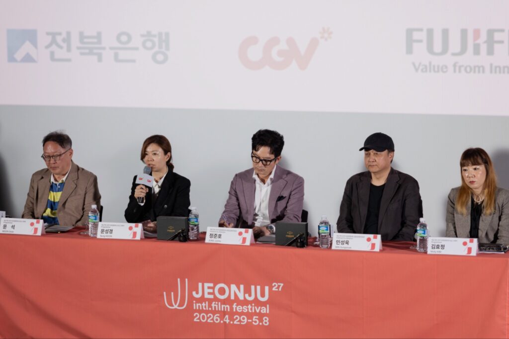 خمسة من المسؤولين والمبرمجين في مهرجان JEONJU International Film يجلسون خلف منصة حمراء خلال المؤتمر الصحفي للإعلان عن النسخة السابعة والعشرين، ويظهر على المنصة شعار المهرجان وتاريخ انعقاده في كوريا الجنوبية.