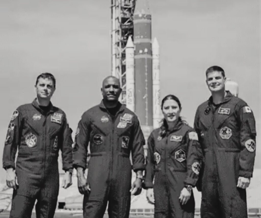 صورة احترافية  بالأبيض والأسود لفريق أرتيميس 2 الأربعة (Reid Wiseman، Victor Glover، Christina Koch، Jeremy Hansen) يقفون معاً ببدلات الطيران الرسمية أمام القمر الكامل ومركبة أوريون، تعبيراً عن المهمة التاريخية التي ستدور حول القمر لمدة ١٠ أيام.