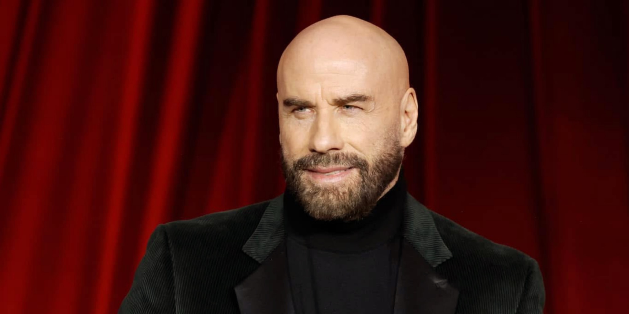 صورة بورتريه مقرّبة ومفصلة للنجم جون ترافولتا John Travolta بأحدث إطلالاته: أصلع، وبذقن كثيفة مشذبة، يرتدي بليزر مخملي أخضر داكن وتيشرت أسود بياقة عالية، يبتسم على خلفية ستائر مسرح حمراء غنية، بمناسبة عودته لمهرجان كان السينمائي 2026.