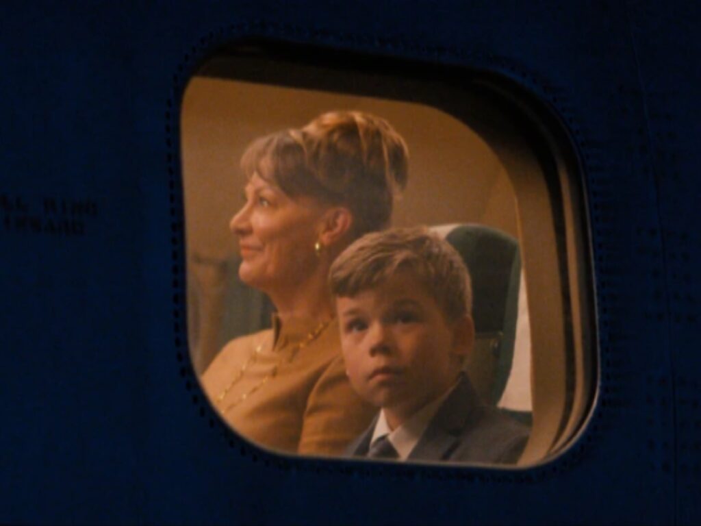 لقطة سينمائية ثابتة، لفيلم  جون ترافولتا تظهر منها نافذة طائرة بيضاوية الشكل. في المقدمة، وولد صغير يرتدي بدلة ورابطة عنق ينظر مباشرة نحو الكاميرا بفضول. في الخلفية، امرأة بالغة بشعر مرفوع بتسريحة عتيقة تنظر بتأمل. كلاهما جالس في مقاعد طائرة كلاسيكية تحت إضاءة دافئة.