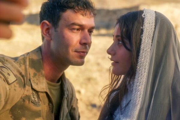 لقطة مقربة (Close-up) من مشهد يظهر الممثل آيتاش شاشماز (Aytaç Şaşmaz) في دور إركان يرتدي زيًا عسكريًا تركيًا مع شارة الهلال والنجمة، ينظر إلى الممثلة هيلين كانديمير (Helin Kandemir) في دور دجلة التي تغطي رأسها بشال أبيض مطرز بخرز دقيق وفريد، كأنهما يلتقطان صورة "سيلفي" (يظهر جزء من يد شخص في المقدمة) في منطقة صحراوية صخرية، بمناسبة مشهد زواجهما المفاجئ في مسلسل أنت من أحببت.