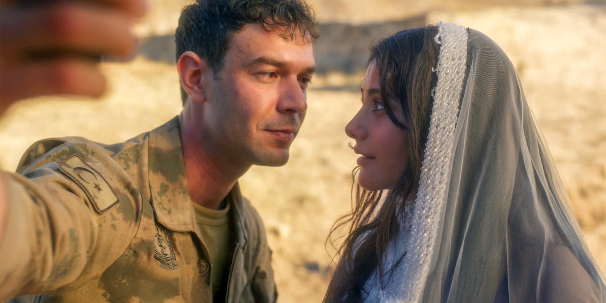 لقطة مقربة (Close-up) من مشهد يظهر الممثل آيتاش شاشماز (Aytaç Şaşmaz) في دور إركان يرتدي زيًا عسكريًا تركيًا مع شارة الهلال والنجمة، ينظر إلى الممثلة هيلين كانديمير (Helin Kandemir) في دور دجلة التي تغطي رأسها بشال أبيض مطرز بخرز دقيق وفريد، كأنهما يلتقطان صورة "سيلفي" (يظهر جزء من يد شخص في المقدمة) في منطقة صحراوية صخرية، بمناسبة مشهد زواجهما المفاجئ في مسلسل أنت من أحببت.