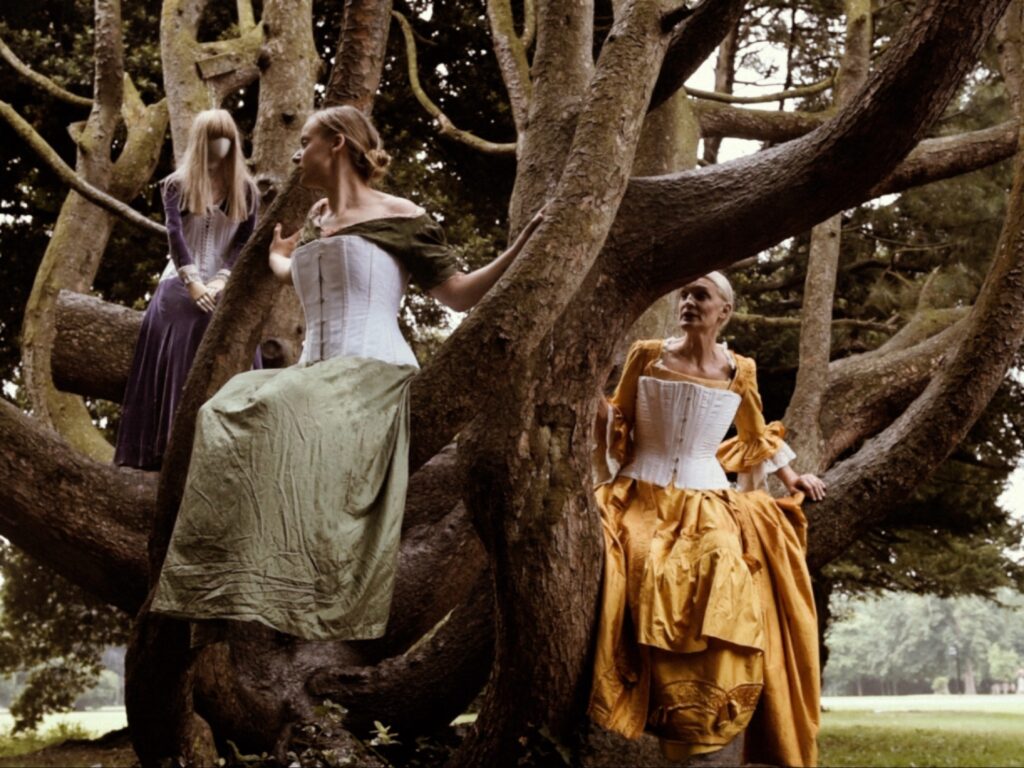 ثلاث شخصيات فوق شجرة: Mannequin، وMillie Daniel-Dempsey، والمخرجة Ingrid Nachstern، يرتدين مشدات وتنانير طويلة.