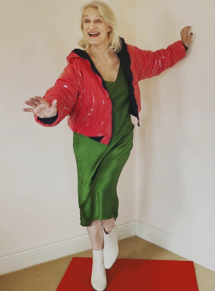 صورة كاملة للمخرجة Ingrid Nachstern تقف متوازنة على قدم واحدة في زاوية غرفة بيضاء، وهي مبتسمة بابتسامة واسعة. ترتدي سترة منفوخة (puffer jacket) باللون الأحمر اللامع وفستاناً حريرياً باللون الأخضر الزمردي، مع بوط بيضاء. يدها اليسرى ممتدة للأمام، واليد اليمنى تستند على الجدار. تقف فوق سجادة حمراء مستطيلة.