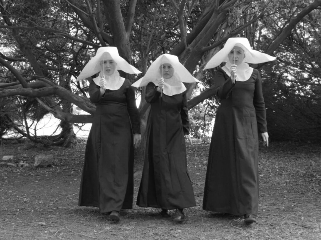 لقطة باللونين الأبيض والأسود لثلاث راهبات يرتدين أغطية رأس بيضاء كبيرة ويسرن للأمام في منطقة مشجرة، وكل منهن تحمل دبوساً رفيعاً أمام وجهها.