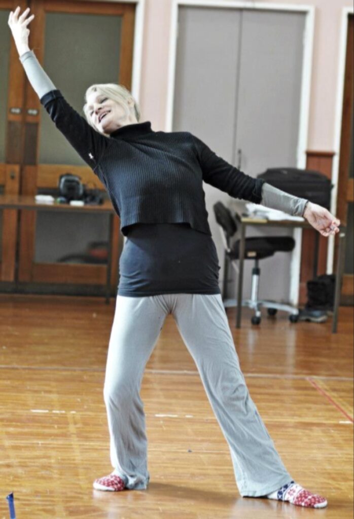 المخرجة Ingrid Nachstern تمارس حركات رقص تعبيري في استوديو، مرتدية ملابس باللونين الأسود والرمادي فوق أرضية خشبية.