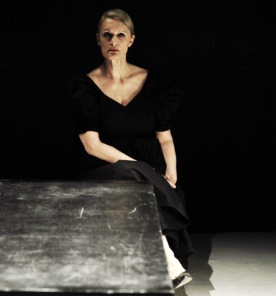 امرأة تجلس خلف طاولة داكنة في غرفة مظلمة، ترتدي فستاناً أسود بأكمام منفوخة وتنظر مباشرة إلى الكاميرا بتعبير جدي تحت إضاءة درامية.