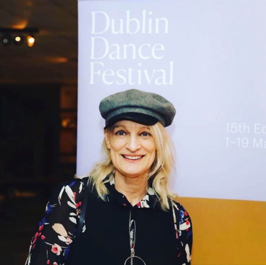 امرأة شقراء مبتسمة ترتدي قبعة رمادية وقميصاً بنقوش ملونة، تقف أمام لافتة مهرجان Dublin Dance Festival.