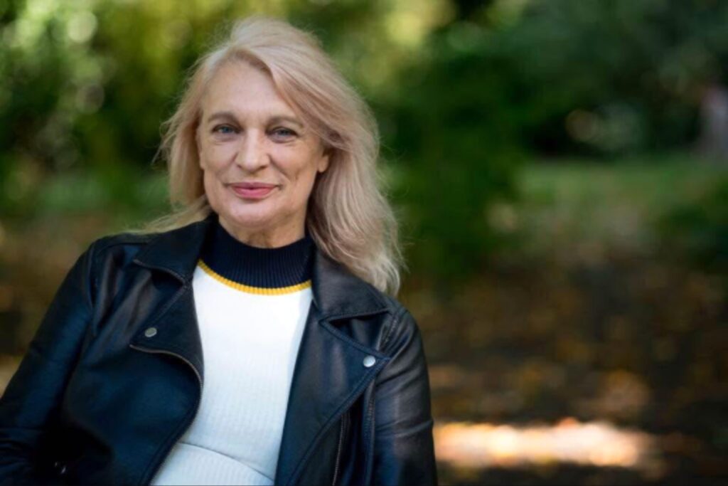 بورتريه لامرأة مبتسمة بملامح هادئة، ترتدي سترة جلدية سوداء فوق قميص أبيض بياقة مخططة، تقف في بيئة خارجية مشجرة بوسط خلفية ضبابية.