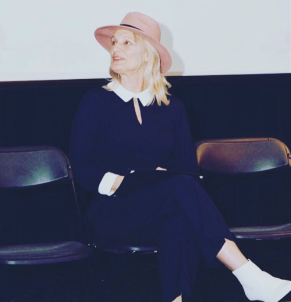 امرأة شقراء تجلس على كرسي أسود، ترتدي زياً كحلياً بياقة بيضاء وقبعة وردية عريضة الحواف وحذاءً أبيض، تنظر إلى الجانب في قاعة ذات خلفية داكنة.