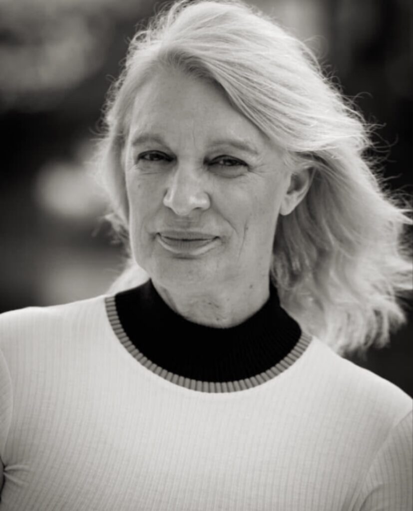 
بورتريه بالأبيض والأسود لامرأة شقراء تنظر مباشرة إلى الكاميرا بملامح هادئة، ترتدي قميصاً فاتح اللون بياقة داكنة عالية، مع خلفية ضبابية.