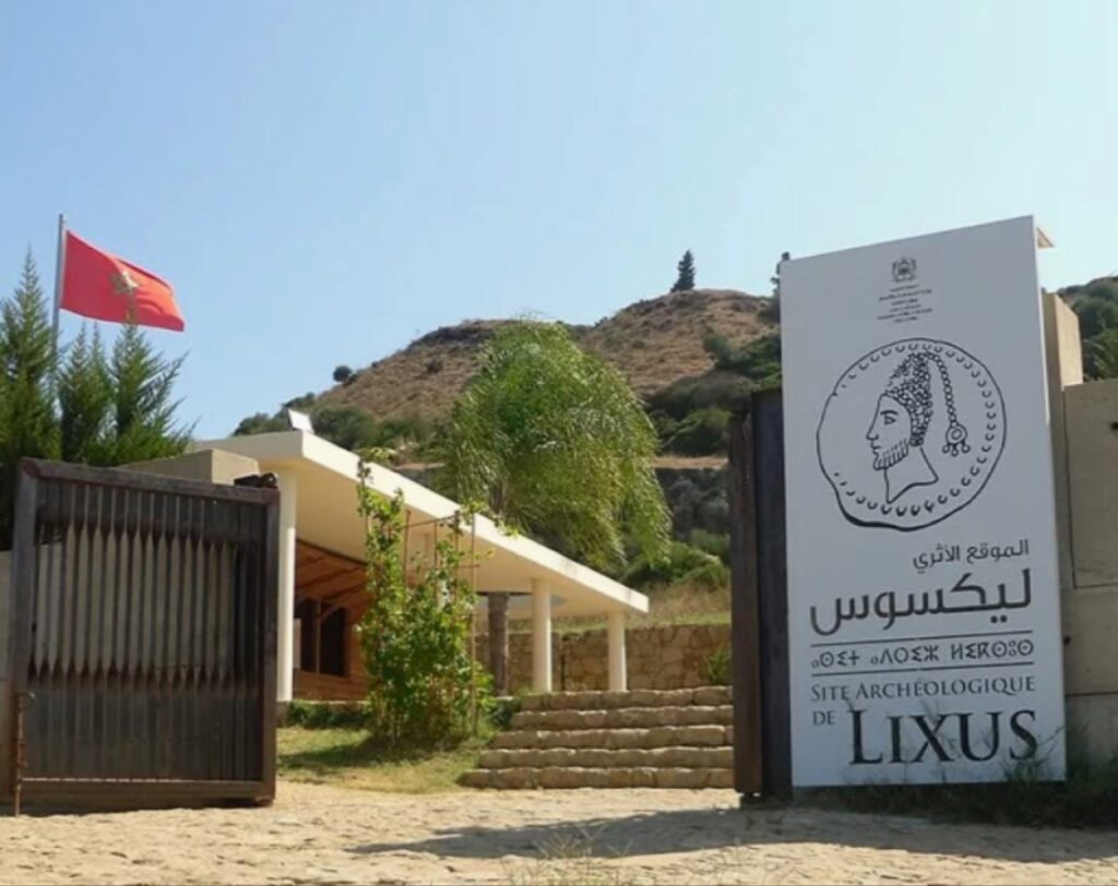 صورة لمدخل "الموقع الأثري ليكسوس" في مدينة العرائش المغربية، يظهر فيها العلم المغربي واللوحة التعريفية الرسمية للموقع، حيث تقام عروض نوستالجيا صدى الأسوار لعام 2026 لإعادة إحياء الذاكرة التاريخية للمكان عبر الفنون والتقنيات الحديثة.