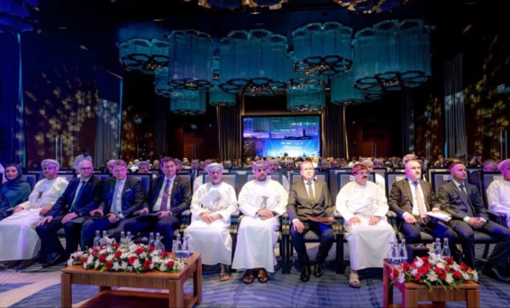 لقطة عامة لقاعة اجتماعات فخمة في مسقط، يظهر فيها صف من كبار المسؤولين ورجال الأعمال العُمانيين والبيلاروس جالسين في الصف الأول يرتدون الثياب التقليدية والبدلات الرسمية، وذلك خلال فعاليات منتدى الأعمال العُماني البيلاروسي لعام 2026، وتتدلى من السقف ثريات زرقاء هندسية مميزة.