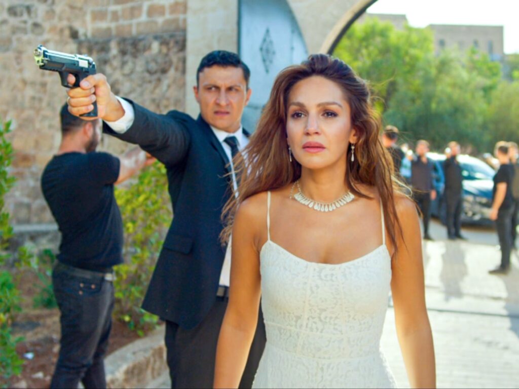 لقطة درامية مليئة بالتوتر من مسلسل المدينة البعيدة، تظهر فيها علياء البورا (سينام أونسال) تسير للأمام بملامح جادة مرتدية فستانًا أبيض من الدانتيل وعقدًا، بينما يقف خلفها جيهان البورا (أوزان أكبابا) ببدلة سوداء رافعًا مسدسًا مصوبًا إياه نحو جانب المشهد لحمايتها، بالقرب من مدخل حجري في يوم مشمس.