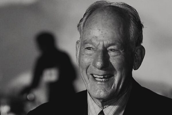 صورة بالأبيض والأسود قريبة للمتسلق جيم ويتاكر يبتسم ويضحك بحرارة. شعره الخفيف مصفف للخلف، ويرتدي سترة داكنة فوق قميص فاتح. تظهر علامات السعادة والشيخوخة على وجهه.