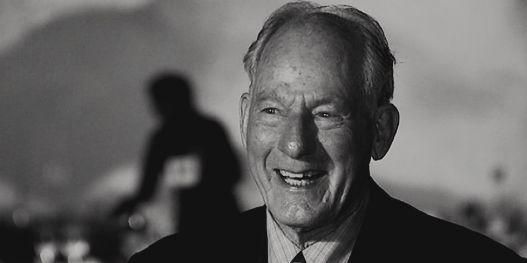 صورة بالأبيض والأسود قريبة للمتسلق جيم ويتاكر يبتسم ويضحك بحرارة. شعره الخفيف مصفف للخلف، ويرتدي سترة داكنة فوق قميص فاتح. تظهر علامات السعادة والشيخوخة على وجهه.