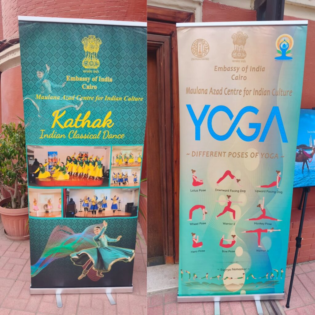 لافتتان (رول أب) واقفتان جنباً إلى جنب أمام مدخل مبنى ضمن فعاليات احتفالية ICCR بالقاهرة. اللافتة اليسرى خضراء اللون، تحمل شعار سفارة الهند بالقاهرة ومركز مولانا آزاد للثقافة الهندية، بعنوان "كاثاك" و"الرقص الكلاسيكي الهندي"، مع صور لراقصات يرتدين أزياء تقليدية. اللافتة اليمنى زرقاء فاتحة، وتحمل نفس الشعارات، بعنوان "يوغا" و"وضعيات مختلفة لليوغا"، مع رسوم توضيحية لعدة وضعيات يوجا كرتونية بما في ذلك لوتس، المحارب 2، ومؤثرات "سوريا ناماسكار".