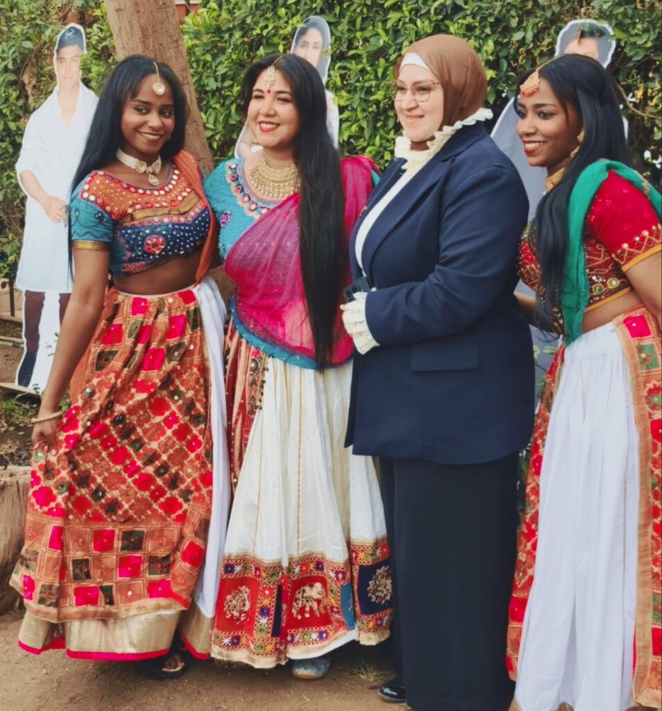 سيدة محجبة ترتدي بدلة زرقاء تلتقط صورة جماعية في حديقة، وتقف بين ثلاث فتيات هنديات يرتدين أزياء هندية تقليدية زاهية ومتنوعة (لِيهينغا)،.