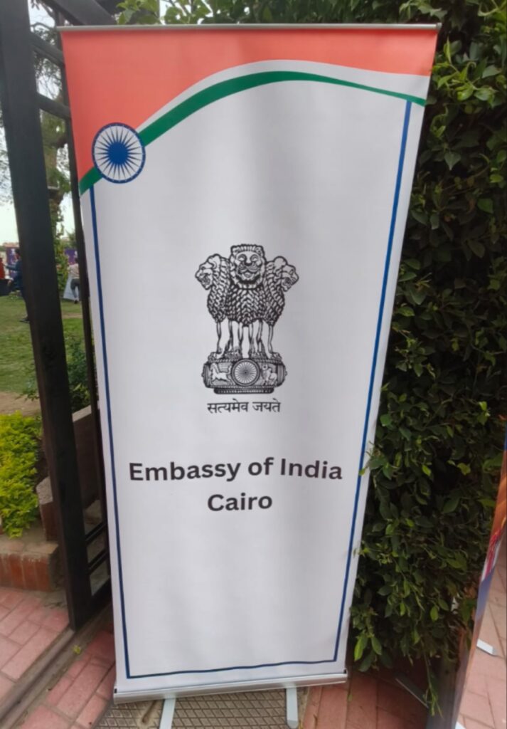 لافتة "رول-آب" بيضاء اللون مع شعار الأسد الهندي الرسمي وشعار سفارة الهند بالقاهرة باللغة الإنجليزية والعربية، تقف أمام شجيرة خضراء.