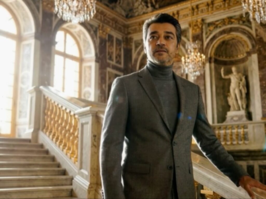 النجم عصام كمال  يرتدي سترة رمادية بياقة عالية يقف على سلم رخامي كبير داخل قاعة فاخرة مزينة بثريات.