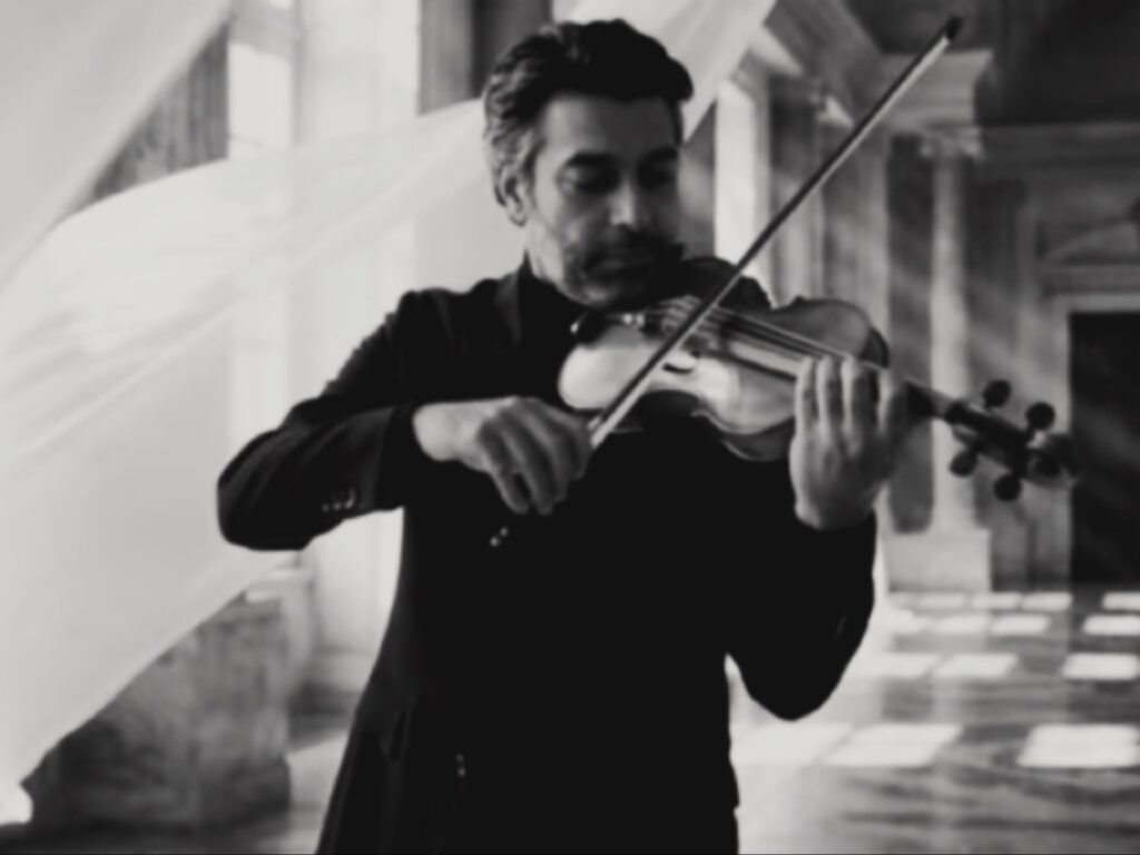 لقطة بالأبيض والأسود لرجل يحمل كماناً ويقوسه بحزن في قاعة فخمة بظلال ضوئية.