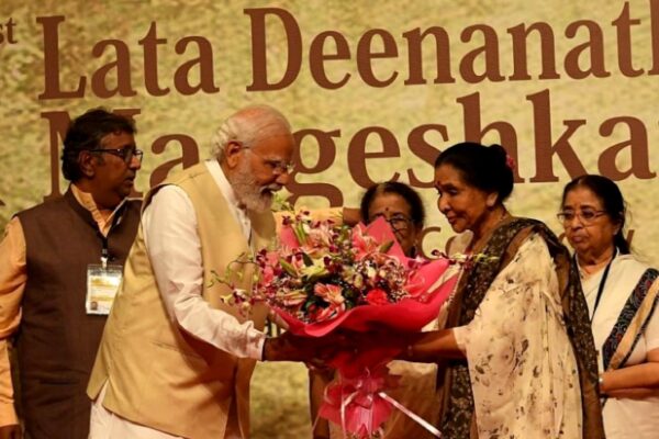 صورة رسمية تظهر رئيس الوزراء الهندي ناريندرا مودي يرتدي صديرياً باللون البيج وهو يهدي باقة ضخمة من الزهور الملونة للفنانة آشا بهوسل التي تطل بسارٍ هندي أبيض بكنار مزخرف. تظهر في الخلفية لافتة كبيرة مكتوب عليها بالإنجليزية "1st Lata Deenanath Mangeshkar" (جائزة لاتا ديناناث منجيشكار الأولى). الإضاءة مسرحية دافئة تعكس أجواء التكريم والوفاء لعام 2026.