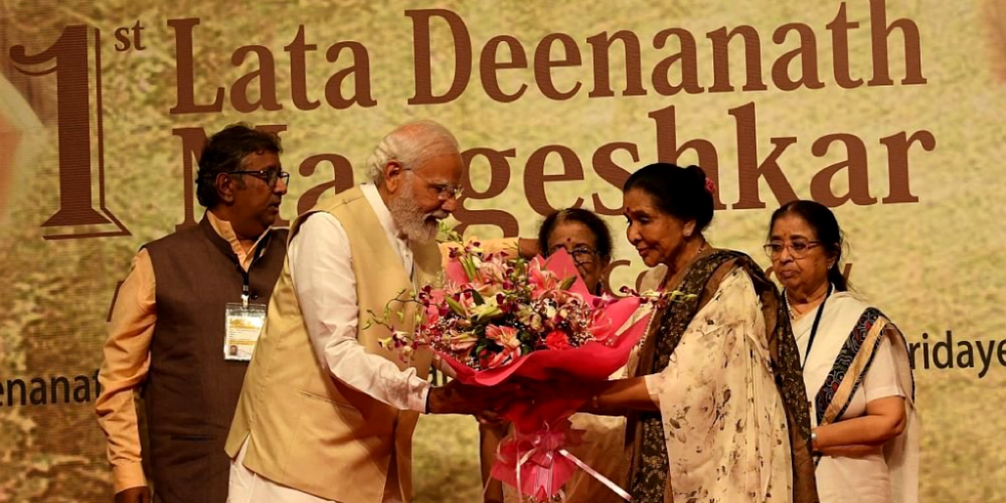 صورة رسمية تظهر رئيس الوزراء الهندي ناريندرا مودي يرتدي صديرياً باللون البيج وهو يهدي باقة ضخمة من الزهور الملونة للفنانة آشا بهوسل التي تطل بسارٍ هندي أبيض بكنار مزخرف. تظهر في الخلفية لافتة كبيرة مكتوب عليها بالإنجليزية "1st Lata Deenanath Mangeshkar" (جائزة لاتا ديناناث منجيشكار الأولى). الإضاءة مسرحية دافئة تعكس أجواء التكريم والوفاء لعام 2026.