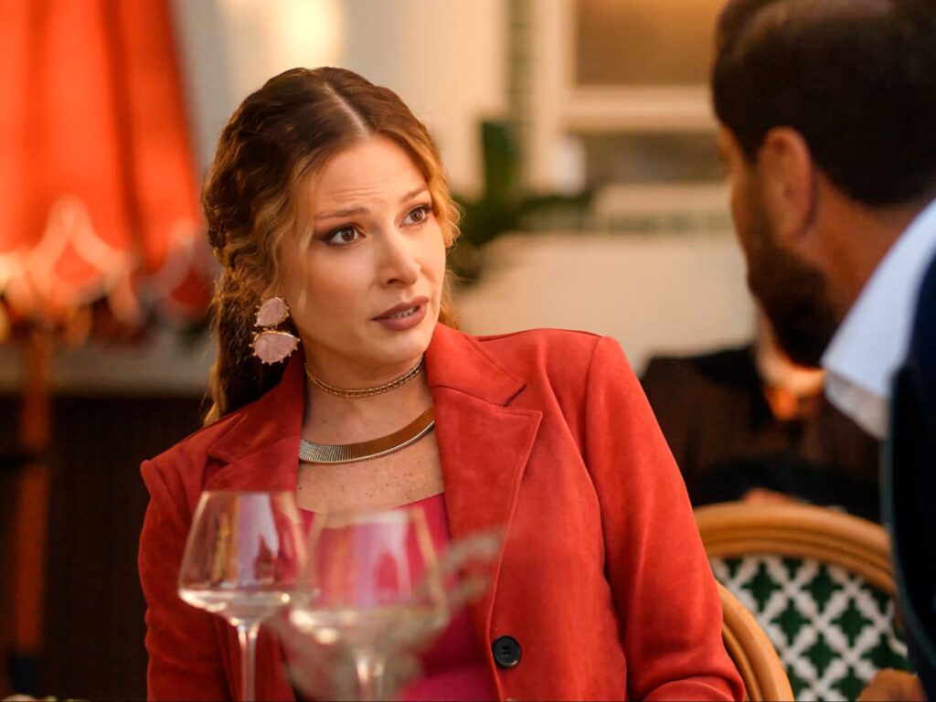 لقطة مقربة لامرأة تتحدث في مطعم، ترتدي سترة حمراء وأقراطاً وردية، مع رجل ينظر إليها على اليمين.