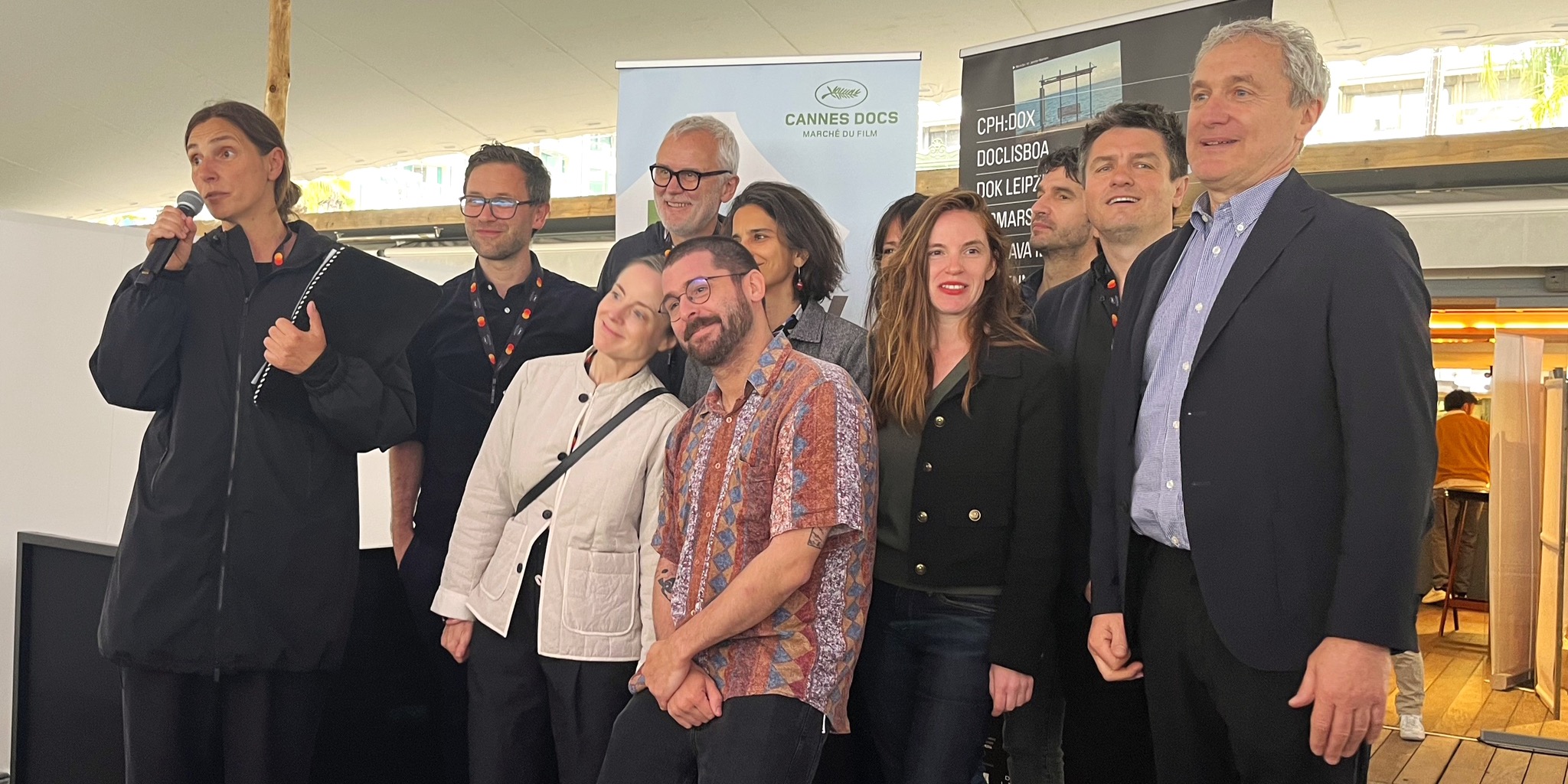 صورة تذكارية لأعضاء تحالف المهرجانات السينمائية الأوروبية (Doc Alliance)، تظهر امرأة تتحدث في ميكروفون ولافتات "DOK Leipzig" و"Marché du Film" خلفهم، خلال حفل مرتبط بـ جائزة Doc Alliance Award 2026.