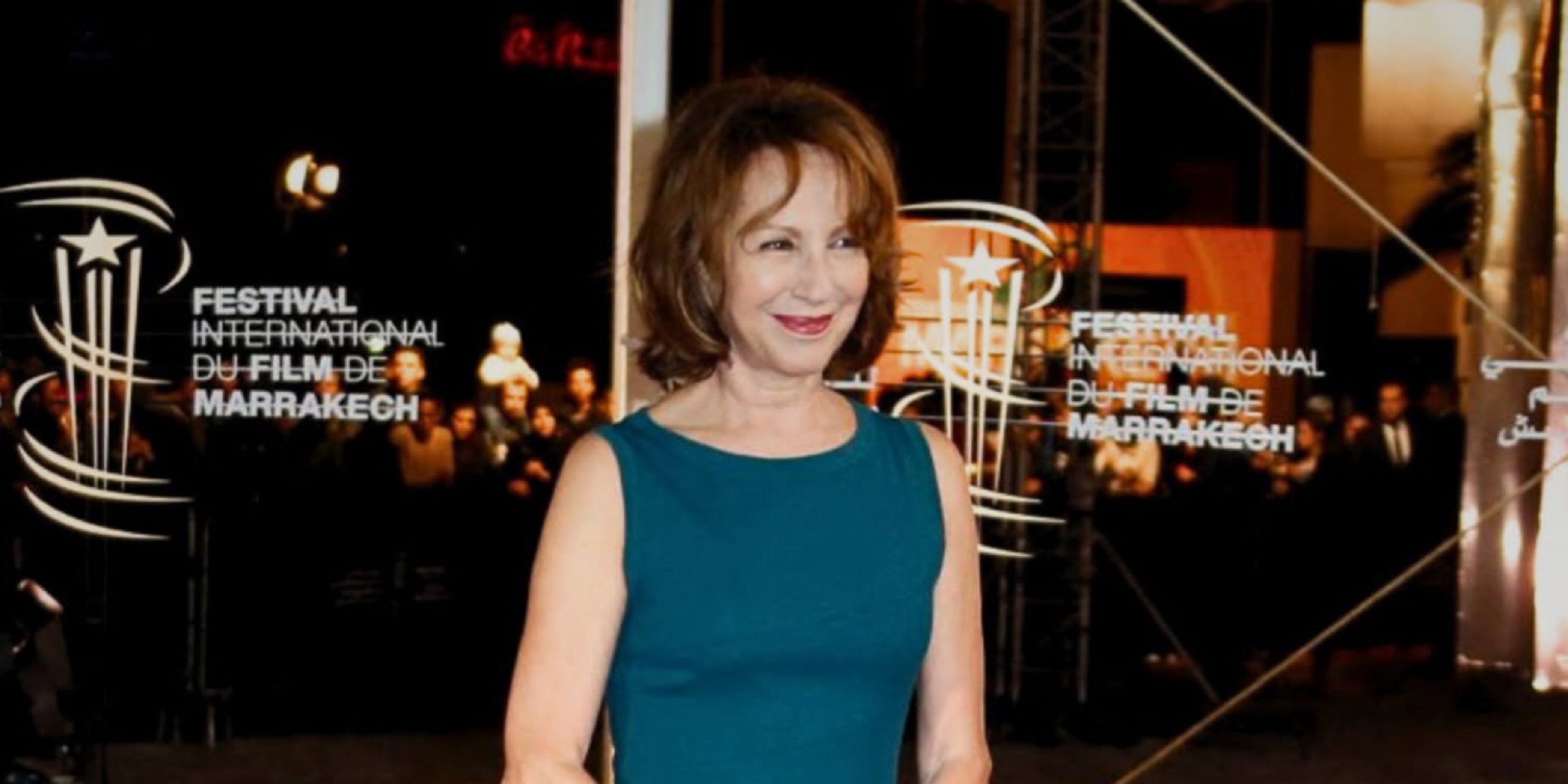 لقطة متوسطة للممثلة ناتالي باي بفستان أزرق داكن، تبتسم وتنظر إلى اليسار، مع ظهور واضح لشعار "FESTIVAL INTERNATIONAL DU FILM DE MARRAKECH" على خلفية مضيئة خلفها.
