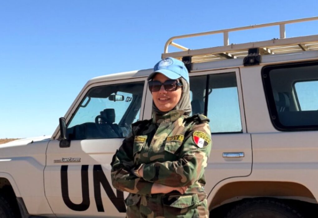 ضابطة مصرية بالزي العسكري المموه وقبعة الأمم المتحدة الزرقاء، تجسد دور مصر بحفظ السلام وتقف أمام مركبة دفع رباعي بيضاء تحمل شعار (UN) في منطقة صحراوية.