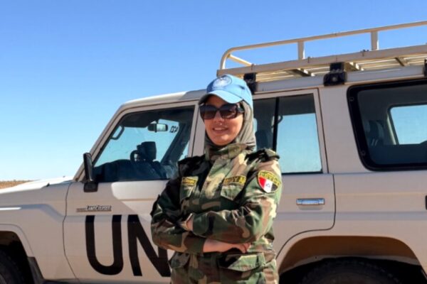 ضابطة مصرية بالزي العسكري المموه وقبعة الأمم المتحدة الزرقاء، تجسد دور مصر بحفظ السلام وتقف أمام مركبة دفع رباعي بيضاء تحمل شعار (UN) في منطقة صحراوية.