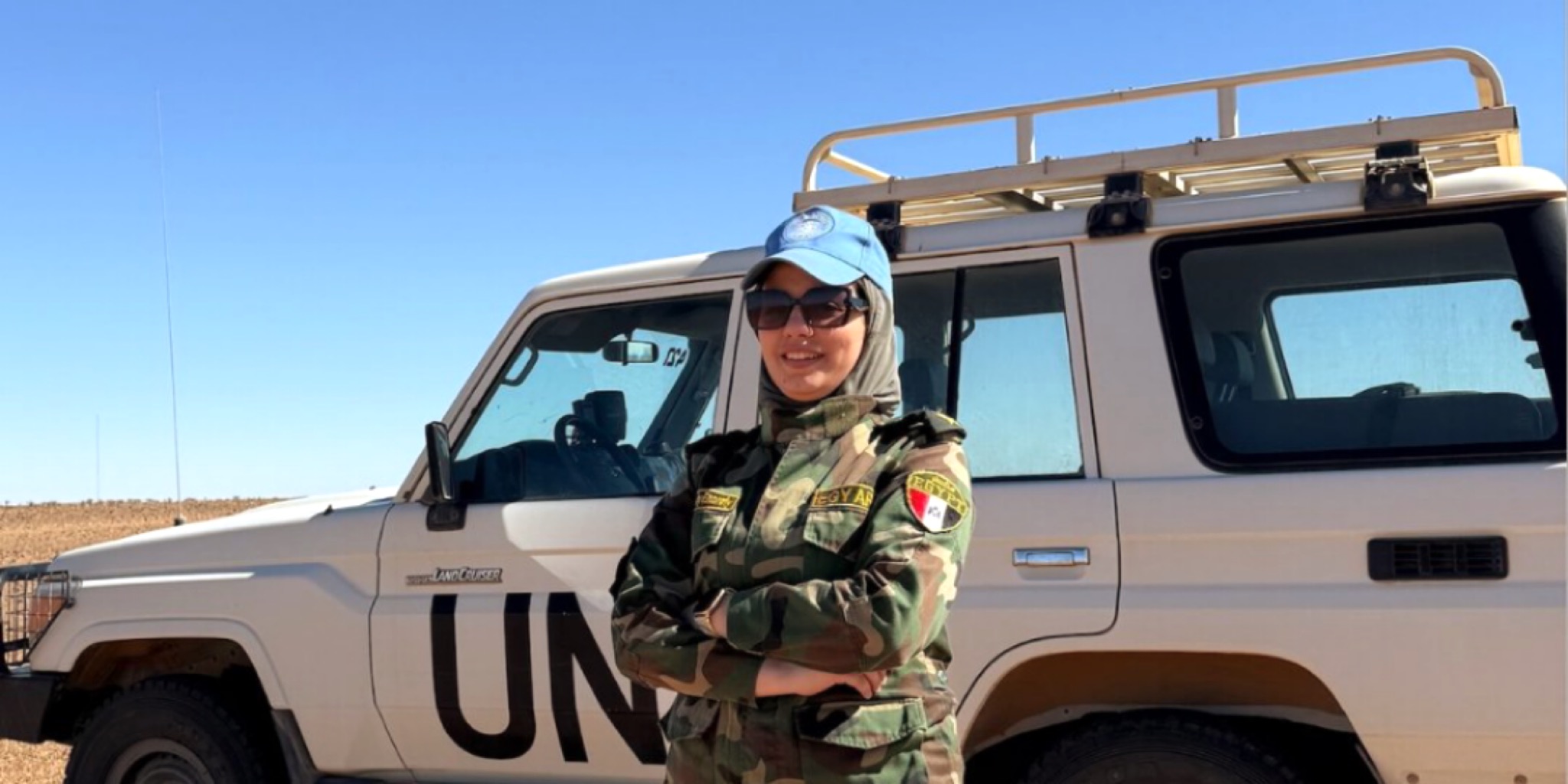 ضابطة مصرية بالزي العسكري المموه وقبعة الأمم المتحدة الزرقاء، تجسد دور مصر بحفظ السلام وتقف أمام مركبة دفع رباعي بيضاء تحمل شعار (UN) في منطقة صحراوية.