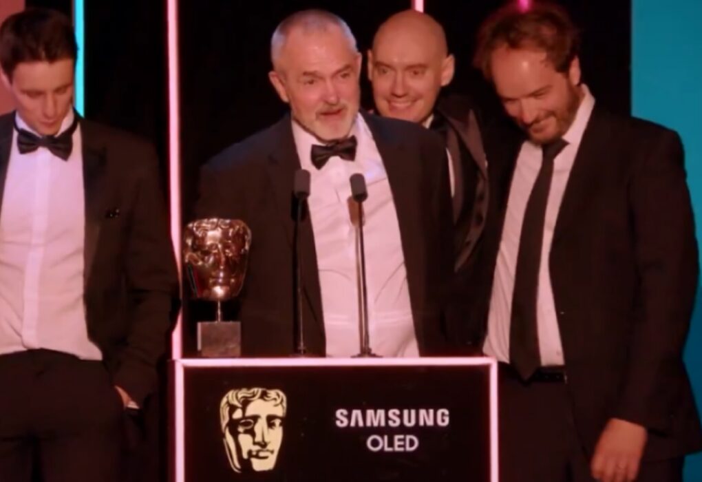 صورة احتفالية لمجموعة من الرجال يرتدون بدلات رسمية "تكسيدو" على المسرح خلف منصة تحمل تمثال بافتا الذهبي وشعار سامسونج OLED، مضاءين بأضواء حفلات الجوائز، يمثلون لحظة الفوز الرئيسية في عام الجوائز بـ جوائز بافتا كرافت للتميز التقني.
