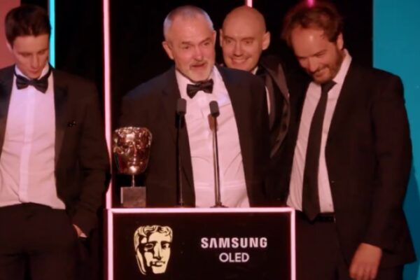 صورة احتفالية لمجموعة من الرجال يرتدون بدلات رسمية "تكسيدو" على المسرح خلف منصة تحمل تمثال بافتا الذهبي وشعار سامسونج OLED، مضاءين بأضواء حفلات الجوائز، يمثلون لحظة الفوز الرئيسية في عام الجوائز بـ جوائز بافتا كرافت للتميز التقني.