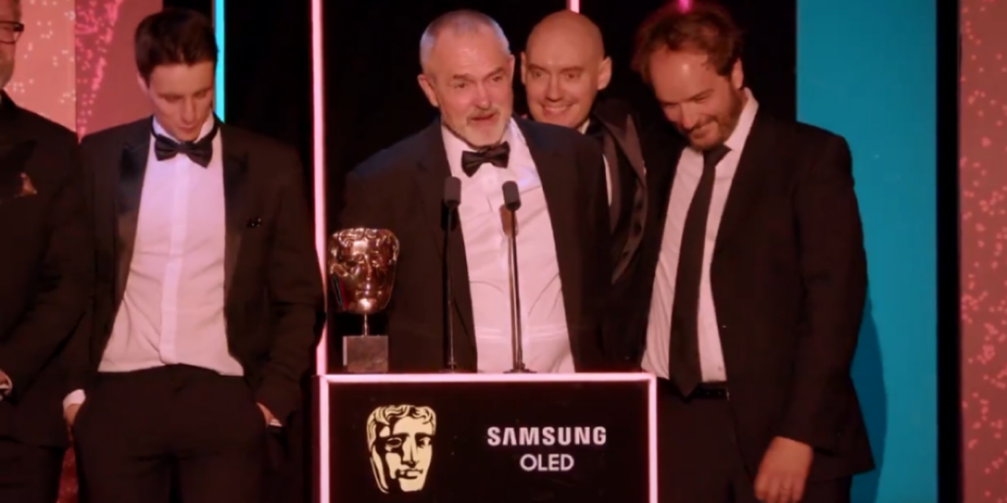 صورة احتفالية لمجموعة من الرجال يرتدون بدلات رسمية "تكسيدو" على المسرح خلف منصة تحمل تمثال بافتا الذهبي وشعار سامسونج OLED، مضاءين بأضواء حفلات الجوائز، يمثلون لحظة الفوز الرئيسية في عام الجوائز بـ جوائز بافتا كرافت للتميز التقني.