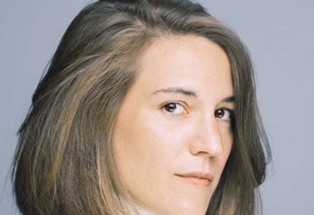 لقطة قريبة لوجه المخرجة الإسبانية كارلا سيمون بنظرة مباشرة وشعر بني منسدل على خلفية رمادية فاتحة.