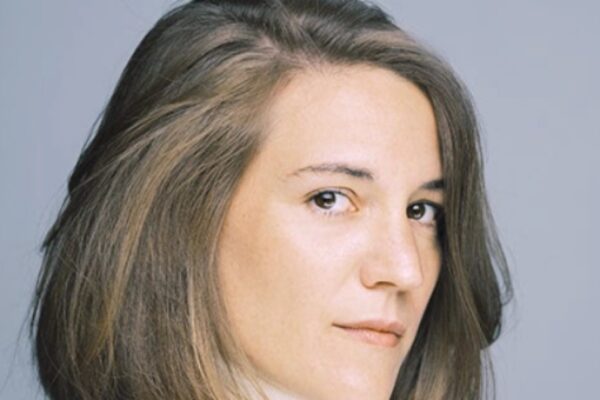 لقطة قريبة لوجه المخرجة الإسبانية كارلا سيمون بنظرة مباشرة وشعر بني منسدل على خلفية رمادية فاتحة.