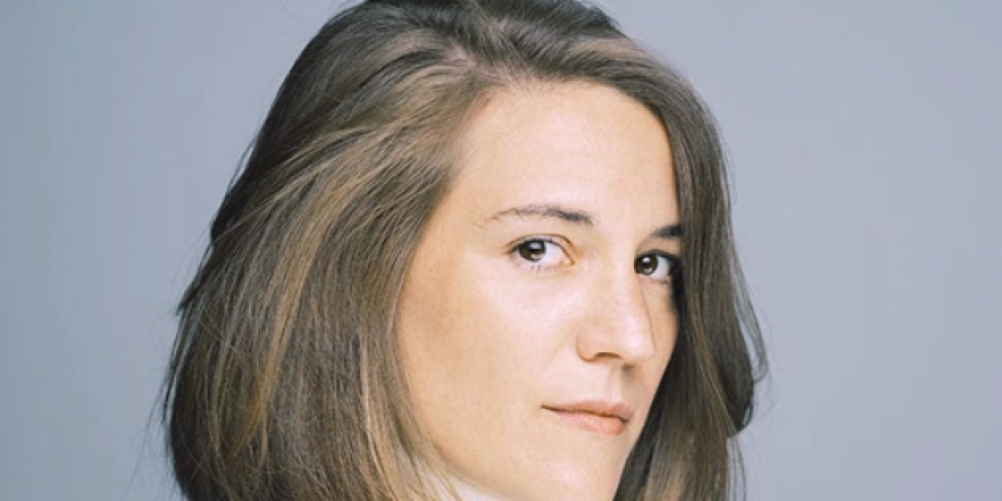 لقطة قريبة لوجه المخرجة الإسبانية كارلا سيمون بنظرة مباشرة وشعر بني منسدل على خلفية رمادية فاتحة.