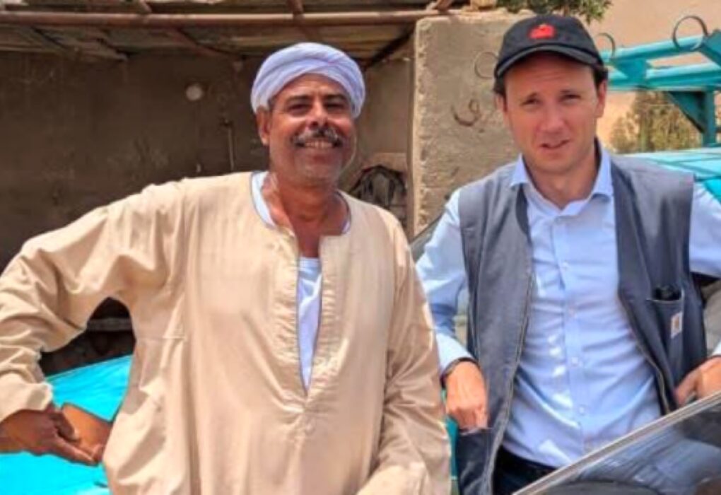 صورة بورتريه تجمع الملحق الثقافي الألماني تيلو فون غيلزا (يمين) يرتدي قبعة بيسبول وسترة، مع مواطن أسواني (يسار) يرتدي الجلباب والعمامة، بجوار سيارة زرقاء، تجسد التبادل بين الثقافة الألمانية والمجتمع المحلي.