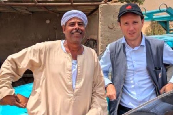 صورة بورتريه تجمع الملحق الثقافي الألماني تيلو فون غيلزا (يمين) يرتدي قبعة بيسبول وسترة، مع مواطن أسواني (يسار) يرتدي الجلباب والعمامة، بجوار سيارة زرقاء، تجسد التبادل بين الثقافة الألمانية والمجتمع المحلي.