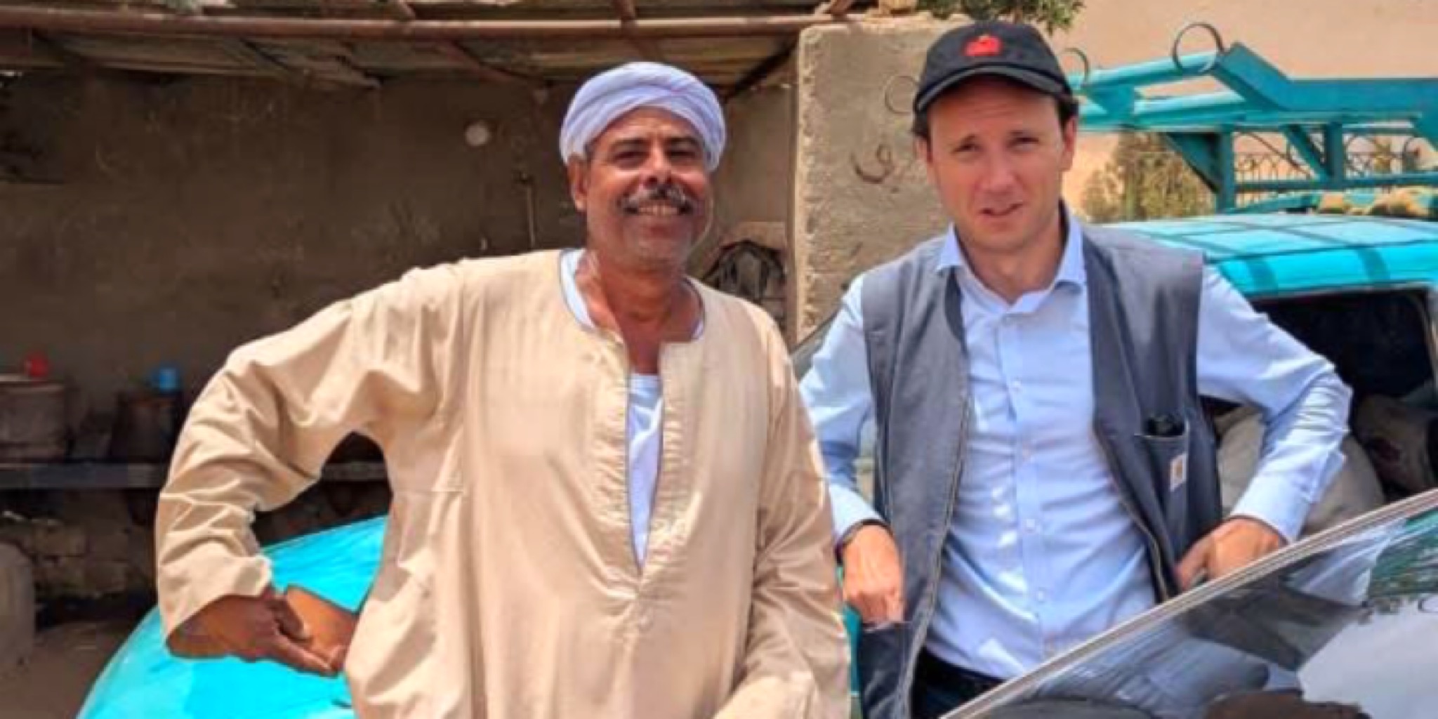 صورة بورتريه تجمع الملحق الثقافي الألماني تيلو فون غيلزا (يمين) يرتدي قبعة بيسبول وسترة، مع مواطن أسواني (يسار) يرتدي الجلباب والعمامة، بجوار سيارة زرقاء، تجسد التبادل بين الثقافة الألمانية والمجتمع المحلي.