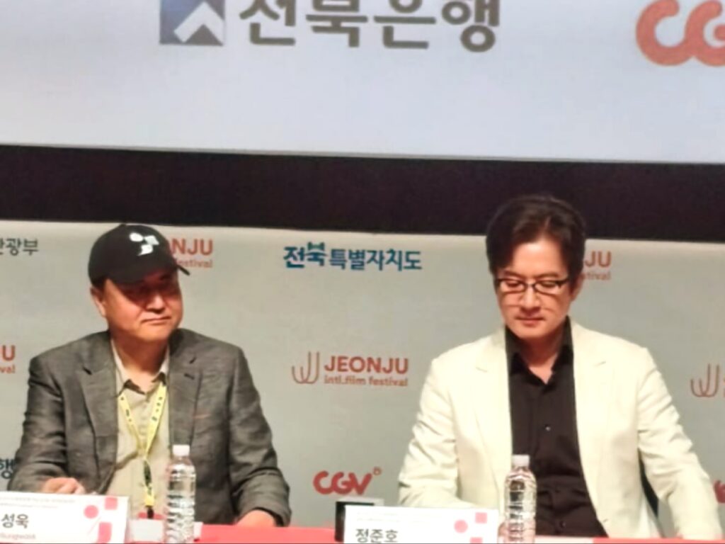 لقطة مقربة للمديرين التنفيذيين المشتركين لمهرجان جونجو السينمائي الدولي (مين سونغ ووك وجونغ جونهو) جالسين في مؤتمر صحفي، أحدهما يرتدي قبعة سوداء والآخر سترة بيضاء، مع لافتة خلفية تحمل شعارات المهرجان والرعاة.