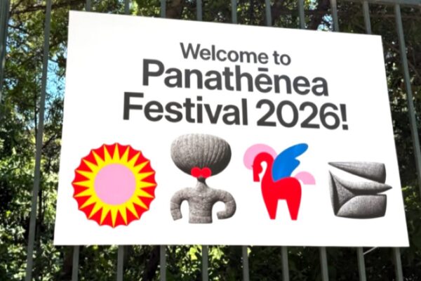 شعار مهرجان باناثينيا 2026 الترحيبي على لافتة بيضاء معلقة وسط الأشجار، يتضمن عبارة Welcome to Panathēnea Festival 2026 والرموز البصرية المميزة للمهرجان