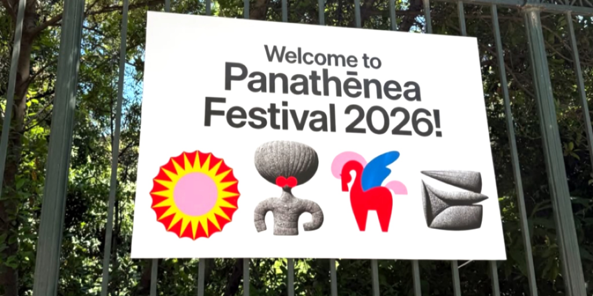 شعار مهرجان باناثينيا 2026 الترحيبي على لافتة بيضاء معلقة وسط الأشجار، يتضمن عبارة Welcome to Panathēnea Festival 2026 والرموز البصرية المميزة للمهرجان