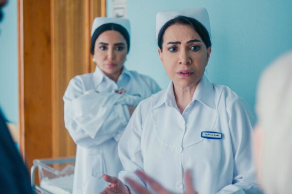 صورة من مسلسل سستر فخرية تظهر النجمة هدى حسين بزي التمريض الأبيض وقبعة الممرضات، تقف في ممر مستشفى بملامح يكسوها القلق والصدمة أثناء حديثها بانفعال مع شخصين، بينما تقف ممرضة أخرى خلفها تتابع الموقف بتوتر