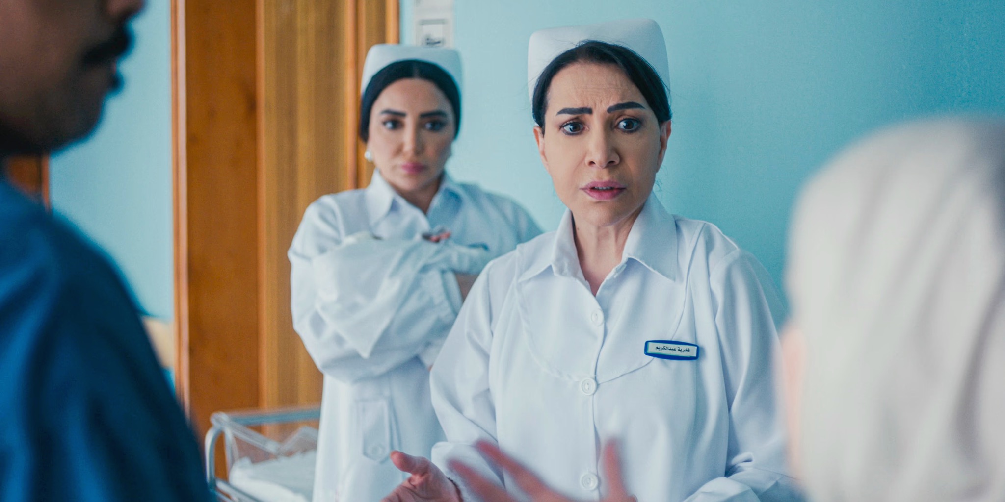 صورة من مسلسل سستر فخرية تظهر النجمة هدى حسين بزي التمريض الأبيض وقبعة الممرضات، تقف في ممر مستشفى بملامح يكسوها القلق والصدمة أثناء حديثها بانفعال مع شخصين، بينما تقف ممرضة أخرى خلفها تتابع الموقف بتوتر