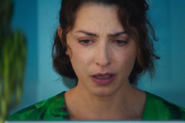لقطة مقربة عاطفية للممثلة باربارا ليني وهي تلعب دور إلسا الباكية في فيلم AMARGA NAVIDAD، ترتدي قميصاً أخضر نابضاً بالحياة أمام خلفية زرقاء ونبات صبار باهت.