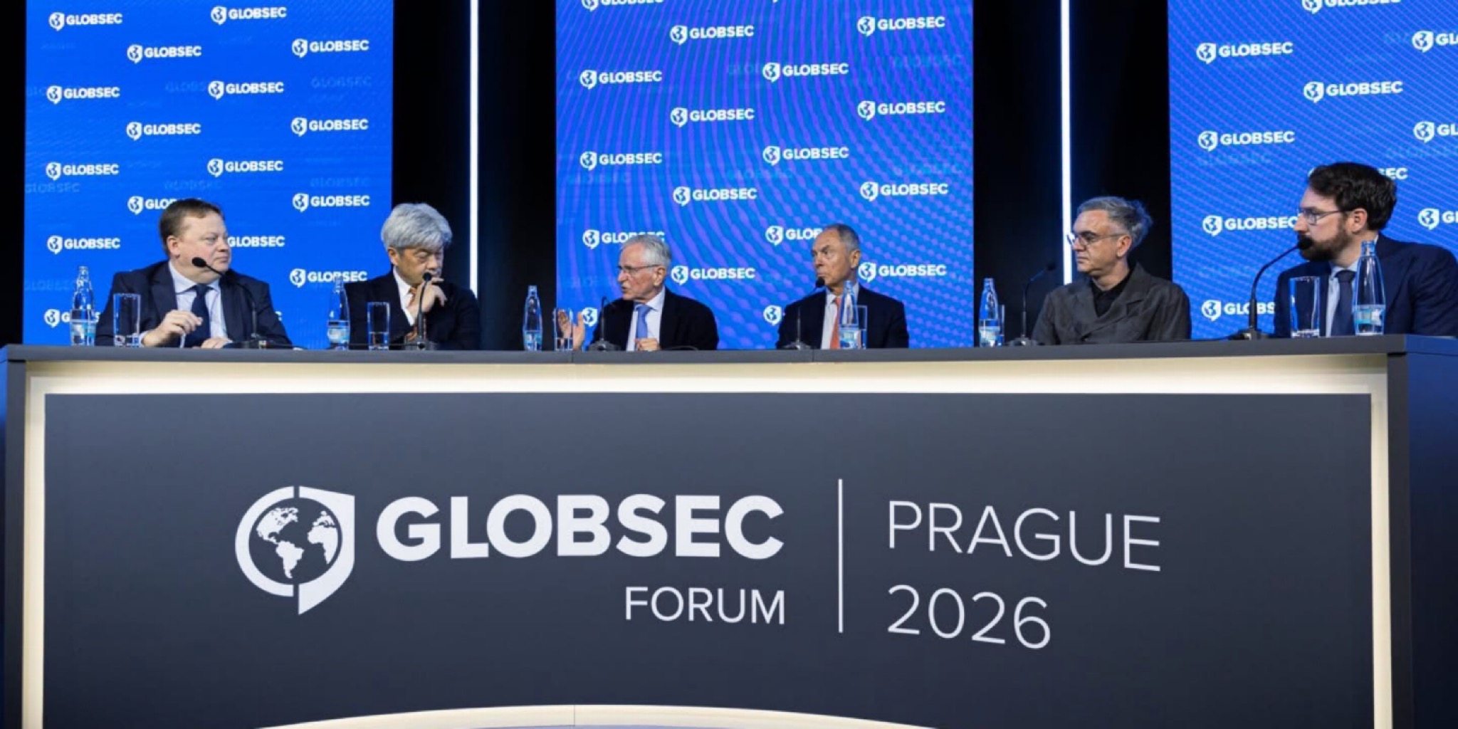 ستة متحدثين يجلسون جنباً إلى جنب خلف طاولة نقاشية عريضة كُتب عليها "GLOBSEC FORUM PRAGUE 2026"، وخلفهم شاشات زرقاء مزينة بشعار غلوبسيك المتكرر، أثناء إحدى الجلسات الحوارية في المؤتمر