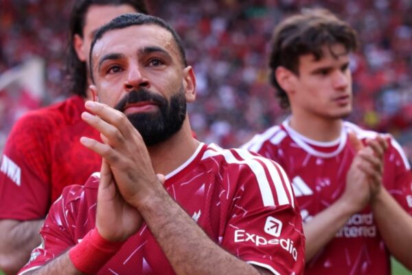 لقطة قريبة توثق وداع محمد صلاح لليفربول، حيث يرتدي القميص الأحمر، يبدو متأثراً بشدة وعيناه تدمعان، وهو يصفق بيديه مودعاً الجماهير في ملعب أنفيلد. يظهر يمين الإطار خلفه لاعب آخر من الفريق، مع خلفية لمدرجات مكتظة بالمشجعين